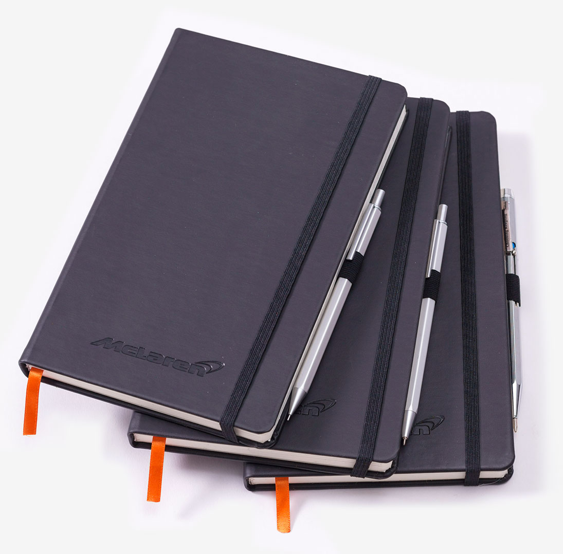 Set of 3 black faux leather notebooks with orange ribbons, black elastic bands and pen loops holding chrome metal pens
