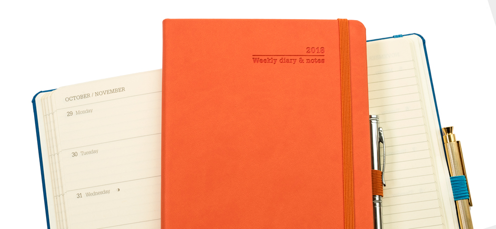 Orange cover 2018 weekly diary with chrome metal pen, and open blue weekly diary with gold metal pen